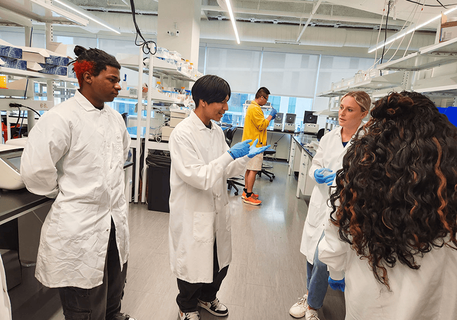 A diverse group of students in labcoats in conversation with mentor inside a lab