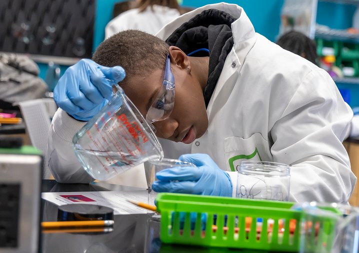 Young male student in lab coat and lab goggles measures liquid with measuring cup